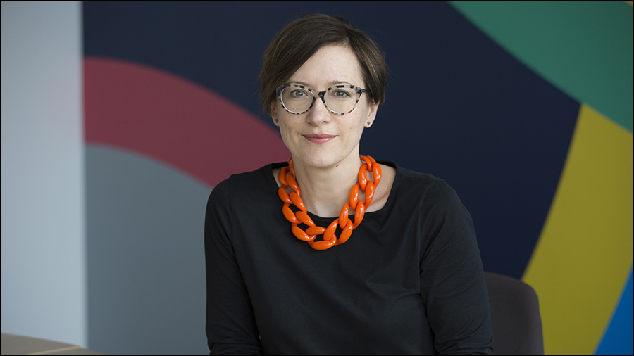 A person with short hair, glasses, and an orange necklace, wearing a black shirt, sits in front of a colorful background. With a keen eye for detail and expertise in HR, they embody the dynamic spirit of people management.