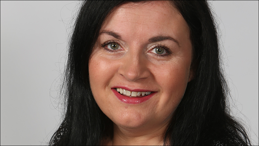 A woman with long black hair and light-colored eyes, possibly an HR professional, smiles at the camera against a plain background.