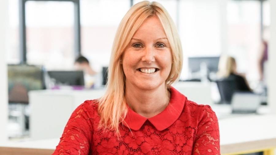 A person with long blonde hair, wearing a red lace top, is smiling in an office setting filled with busy personnel working in the background.