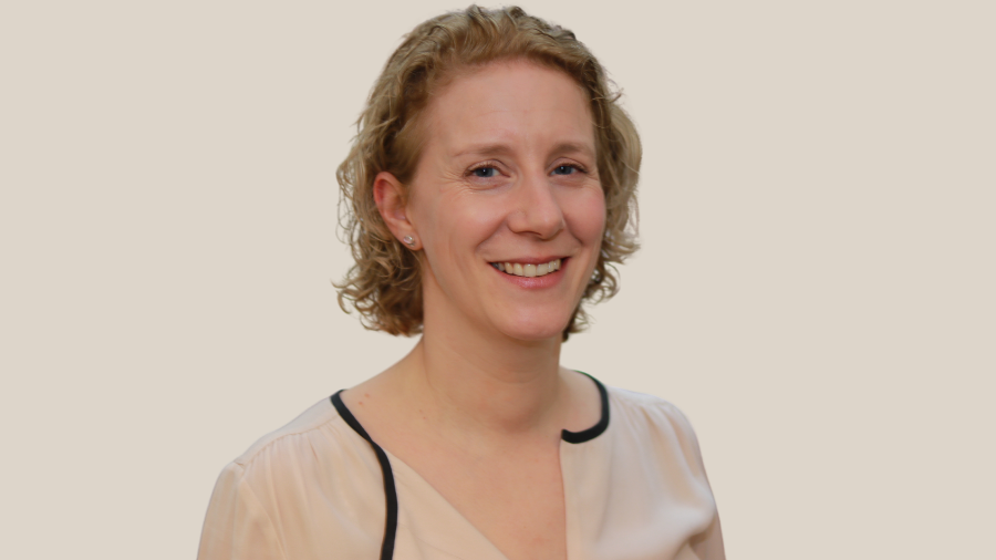 A curly-haired individual wearing a beige blouse with black trim stands against a plain background, smiling at the camera, appearing relieved after receiving their compulsory vaccine.