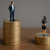 Figurines of a man and woman standing on top of a stack of British coins, the man's taller, demonstrating the gender pay gap.