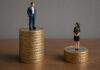 Gender pay gap in tech widens to nine-year high as AI roles drive salaries Figurines of a man and woman standing on top of a stack of British coins, the man's taller, demonstrating the gender pay gap.