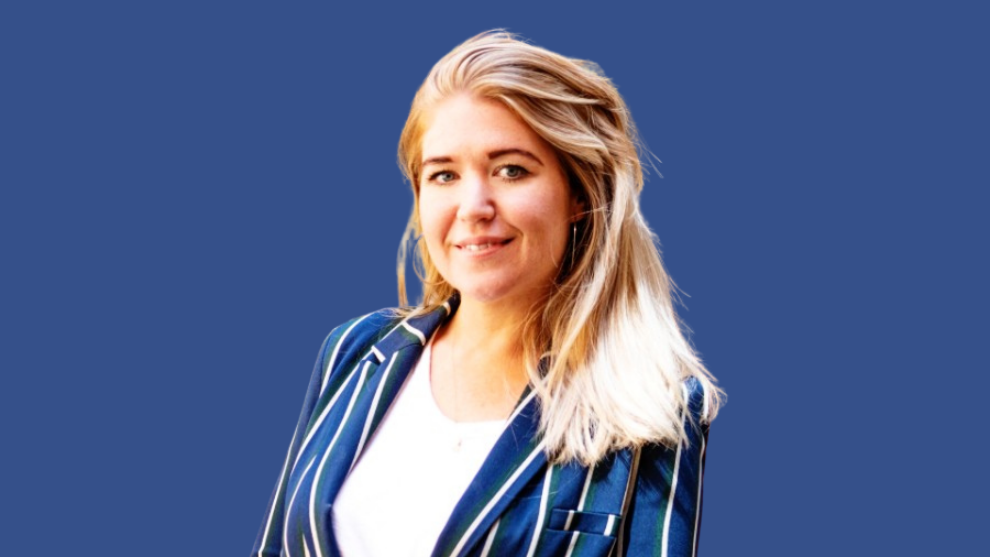 A person with blonde hair, dressed in a striped blazer over a white top, stands against a plain blue background, looking ready to work in the office.