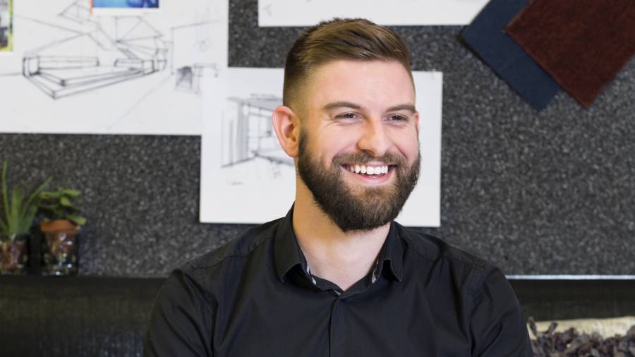 A man with a beard and short hair, wearing a black shirt, is smiling while seated in an office. Sketches and materials are visible on the wall behind him, hinting at his role in People Management.