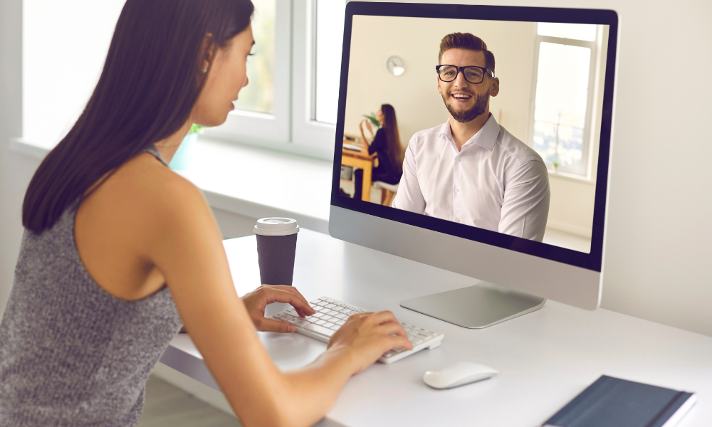 A woman sits at a desk typing on a keyboard, engaged in a video call with a smiling man on her computer screen, discussing HR salaries.
