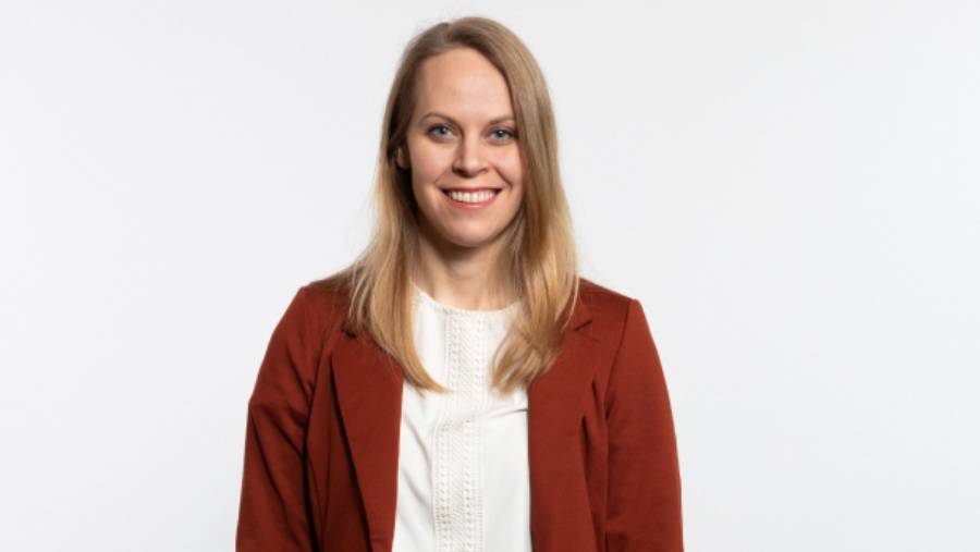 A woman with long blonde hair, wearing a white top and a rust-colored blazer, stands against a plain white background, smiling at the camera. Her confident demeanor suggests she excels in People Management.