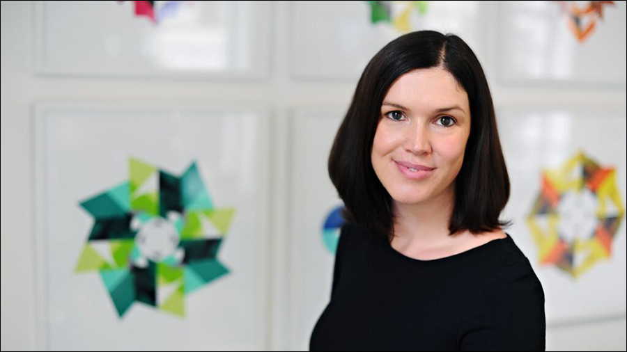 A woman with shoulder-length dark hair and a black top smiles at the camera. Colorful geometric artwork is visible on the wall behind her, creating a vibrant backdrop indicative of an office focused on personnel and people management.