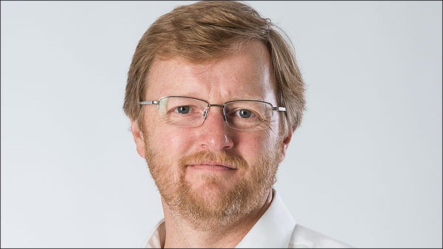 A man with short, light brown hair and a beard is wearing glasses and a white shirt, looking directly at the camera with a neutral expression. With his professional appearance, he seems like someone who excels in Human Resources or personnel management. The background is plain and light-colored.
