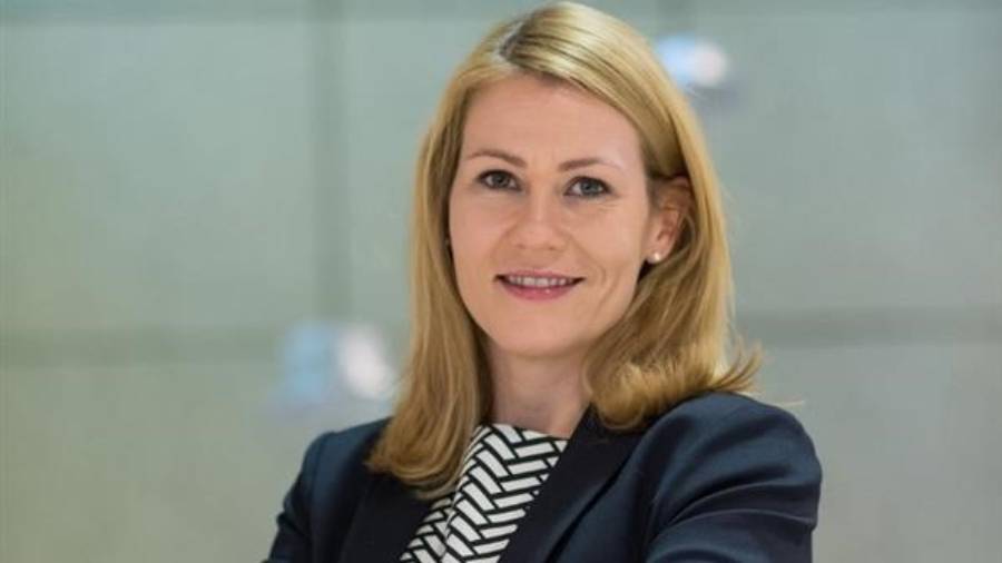 A woman with shoulder-length blonde hair, working in Human Resources, smiles at the camera. She wears a black blazer over a striped shirt. The background is blurred and neutral-toned.