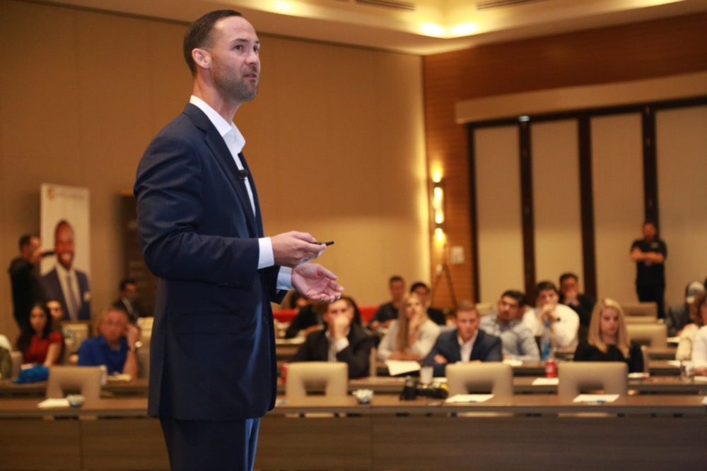A man in a suit gives a presentation to an attentive audience in a conference room, aiming to upskill the professionals. The audience members sit at tables with laptops and other materials.