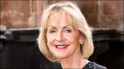 A smiling woman with blonde hair and red lipstick, wearing a dark top with a light pattern, stands in front of a blurred dark background, embodying the essence of people management.