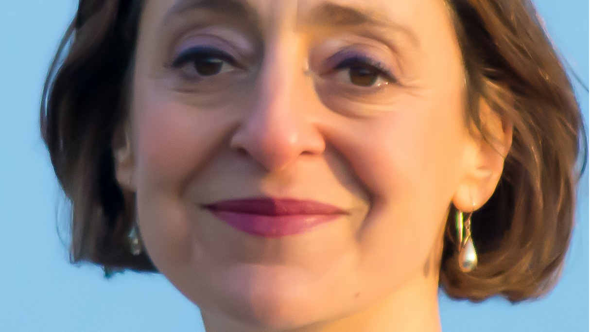 Close-up of a woman with short brown hair, wearing pearl earrings and light purple eyeshadow, smiling slightly with a blue sky in the background—an approachable face that could belong to someone in Human Resources.