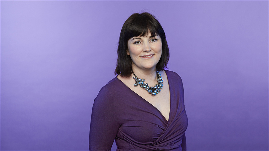 A woman with black hair, wearing a purple dress and a large pearl necklace, stands in front of a purple background, smiling confidently – the epitome of excellence in human resources.