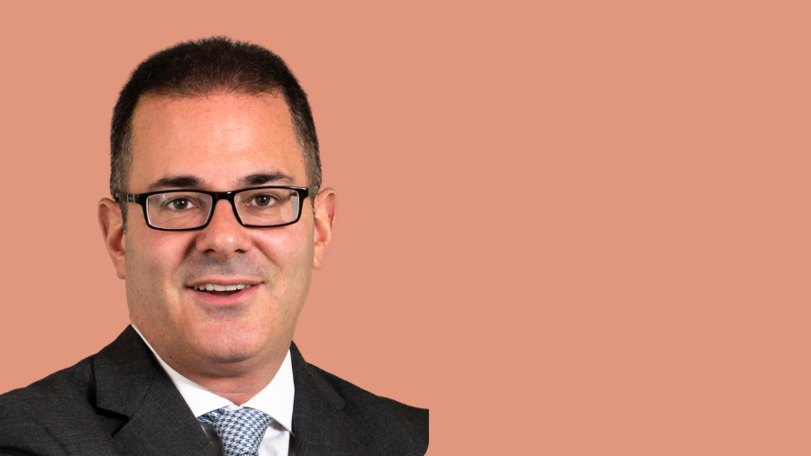 A man with short dark hair and glasses, wearing a grey suit and a light blue tie, smiles against a solid light brown background, embodying the professionalism often seen in Plan B employees.