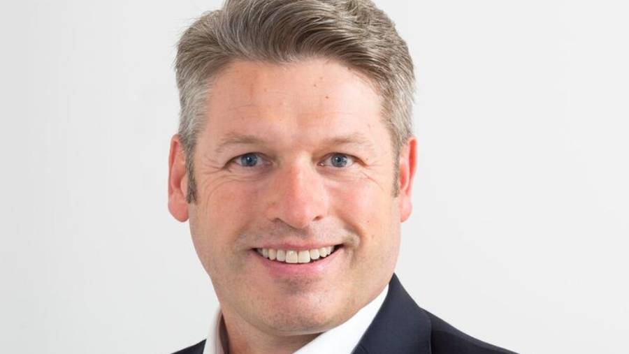 A smiling man with short, light-colored hair, wearing a dark suit jacket, white shirt, and tie, portrays a professional touch in the field of Human Resources against a plain white background.
