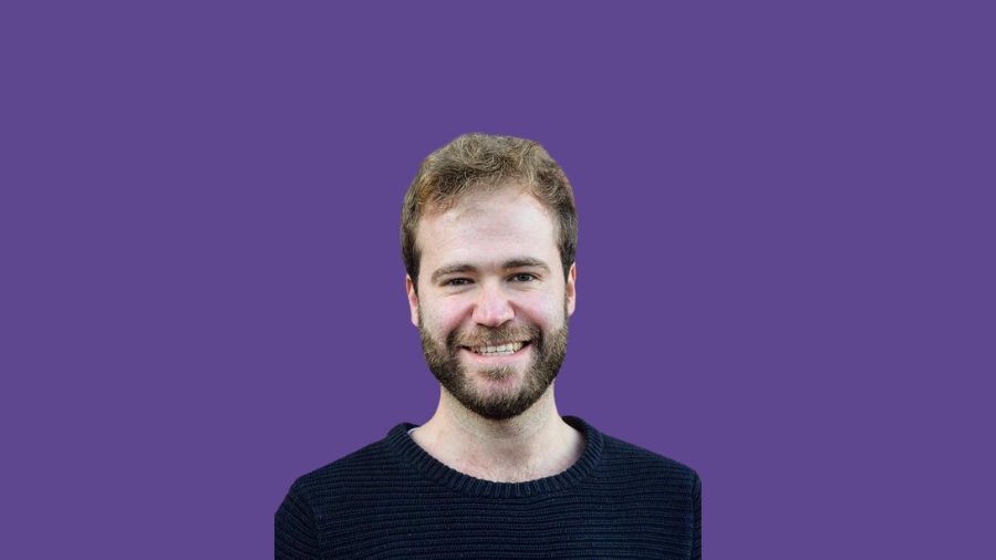 A man with a beard and short hair, wearing a black sweater, smiles in front of a solid purple background, embodying a sense of wellbeing at work. A man with a beard and short hair, wearing a black sweater, smiles in front of a solid purple background, embodying a sense of wellbeing at work.