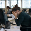 A woman sits at an office desk with her head in her hands.