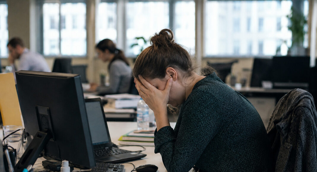 A woman sits at an office desk with her head in her hands.