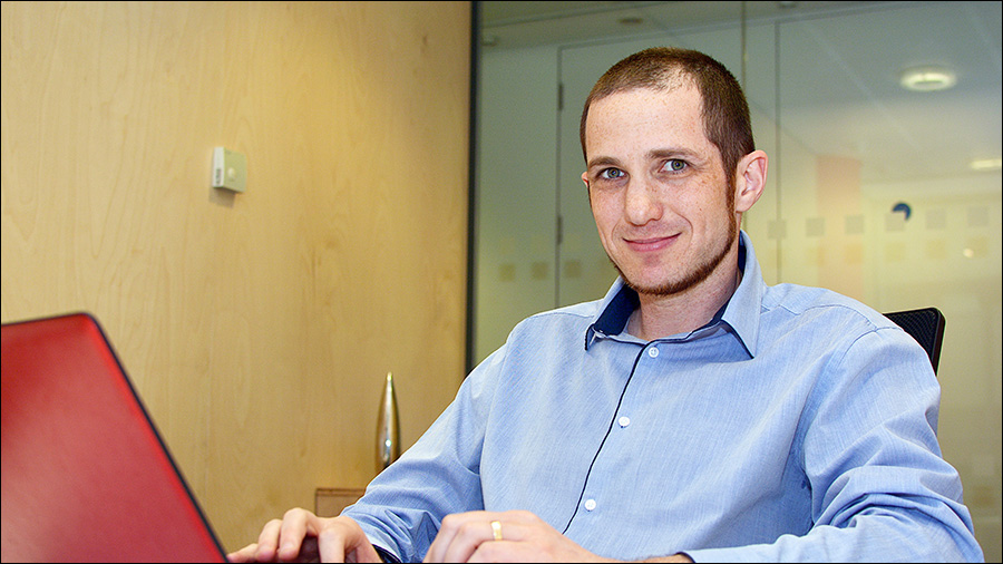 A person wearing a blue shirt is seated at a desk in the Human Resources office, working on a red laptop, smiling while looking at the camera.