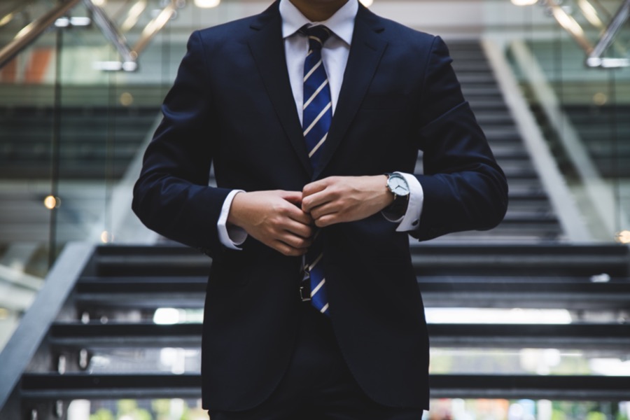 A self-employed individual in a black suit and striped tie stands in front of stairs while buttoning their jacket.