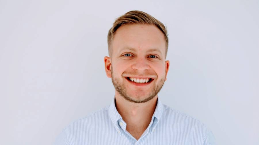 A man with short hair and a beard smiles while wearing a light blue striped shirt, evoking an approachable demeanor suitable for personnel roles or people management, set against a plain white background.