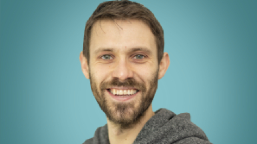 A man with short brown hair and a beard smiles at the camera against a blue background. He is wearing a gray hoodie, radiating the approachable charm of an HR professional.