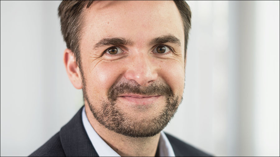 A man with short brown hair and a beard smiles at the camera. He is wearing a white shirt and a dark blazer, exuding the confidence of someone well-versed in people management.