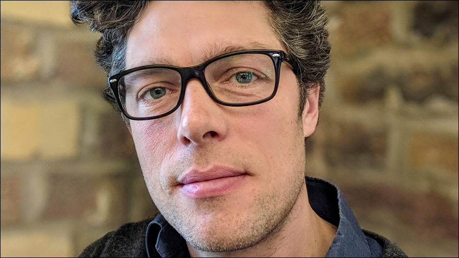 A man with short, wavy hair, wearing black-rimmed glasses and a blue shirt, looks directly at the camera with a neutral expression. The background consists of a blurred brick wall, hinting at his role in Human Resources or People Management.