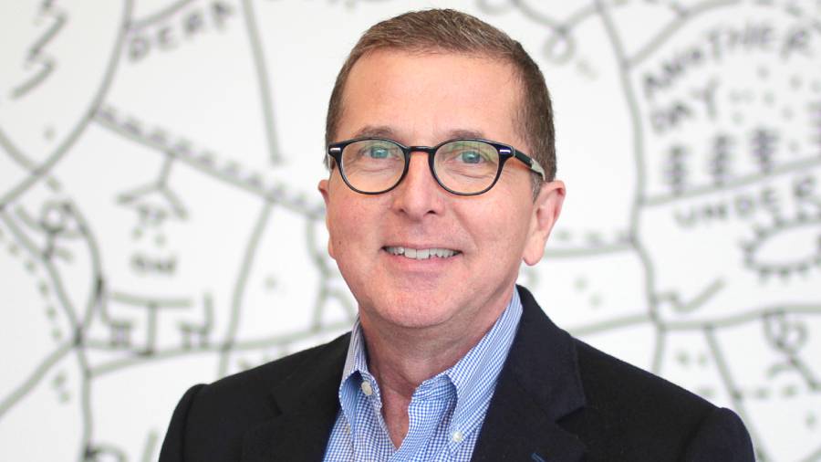 A man wearing glasses and a dark blazer smiles in front of a white wall adorned with a detailed map design, embodying the essence of effective People Management.