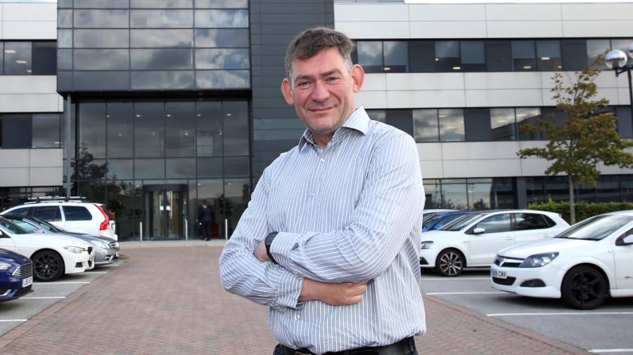 A man in a striped shirt, possibly from Human Resources, stands with folded arms in a parking lot in front of a modern building on a cloudy day. Several cars are parked in the background.