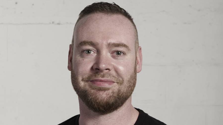 A person with a short haircut and beard, wearing a black shirt, stands in front of a plain white wall, looking at the camera with the poised demeanor of an HR professional.