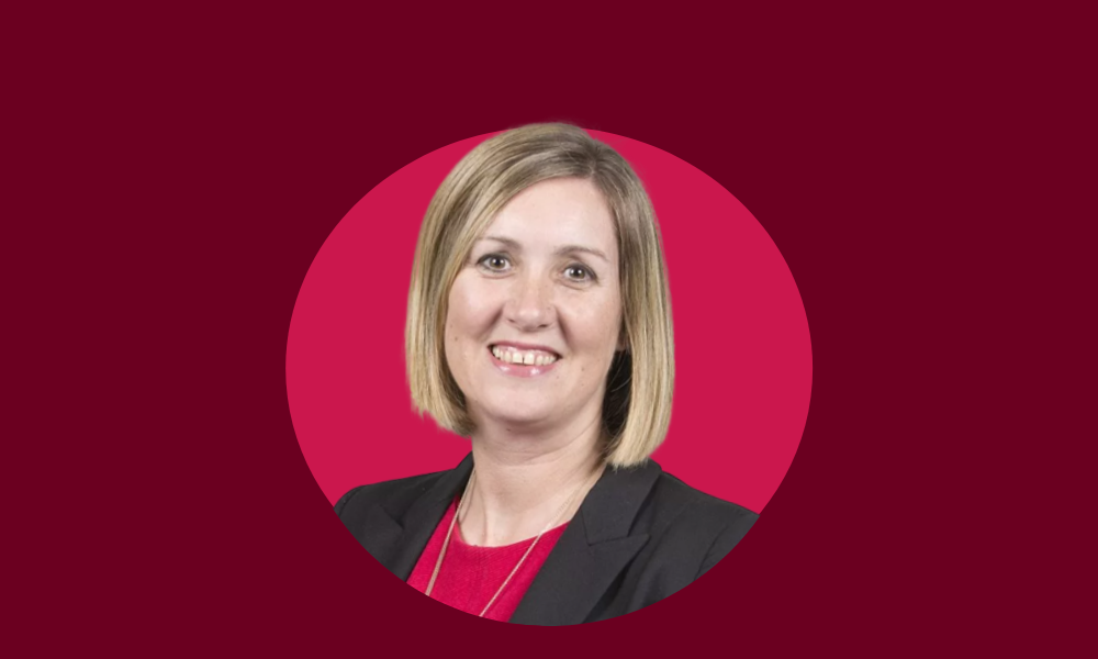 A woman with shoulder-length blonde hair is smiling. She is wearing a black blazer over a red top. The red background subtly highlights the silent strength she carries in her quest against gender inequality.