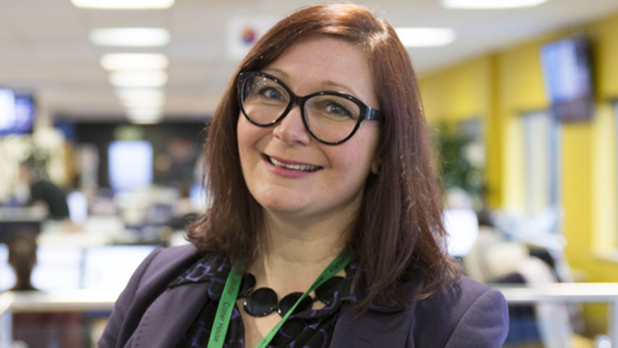 A person with shoulder-length brown hair, wearing glasses, a dark blazer, and a green lanyard, stands smiling in a well-lit Human Resources office space with yellow walls and open-plan workstations.