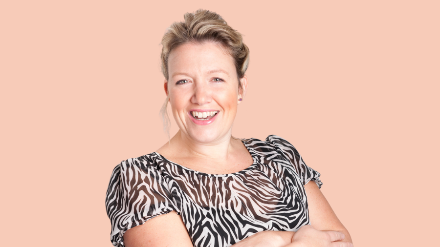 A woman with blonde hair, wearing a black and white zebra print blouse, smiles with her arms crossed in front of a peach-colored background, embodying the spirit of diversity and inclusion.