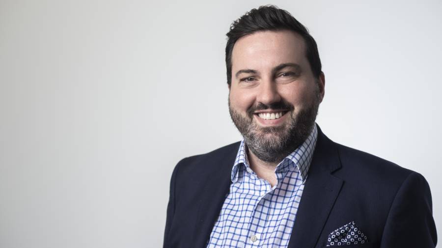 A man with a beard and short dark hair in a blue blazer and checkered shirt smiles against a plain background, embodying the essence of effective People Management.