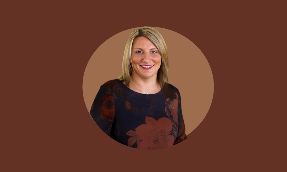 Blonde woman with a smile, wearing a dark floral blouse, standing in front of a circular maroon background. #breakthebias