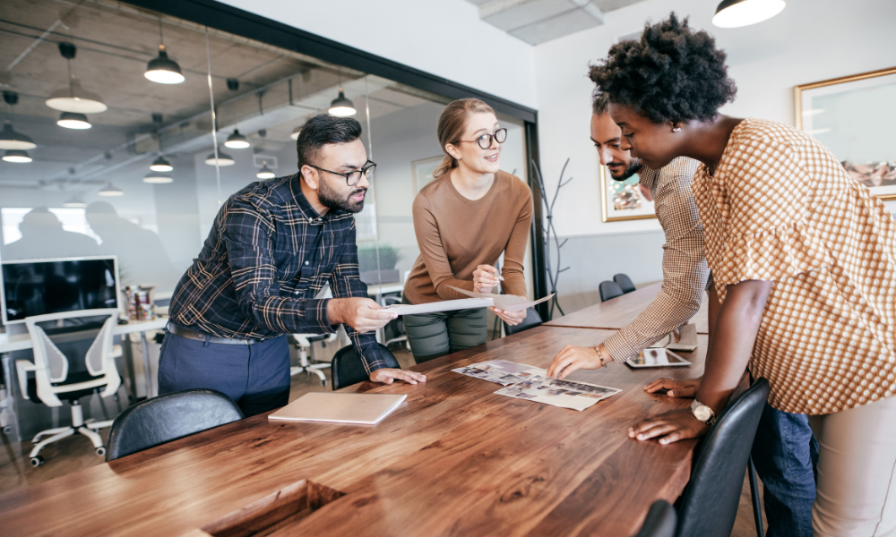 Four UK workers stand around a conference table in an office, reviewing documents and discussing work, mindful of the increasing cost of living.