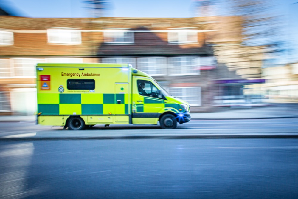 A yellow and green emergency ambulance speeds down a city street with buildings in the background, captured with motion blur, emphasizing the urgency of health and safety.