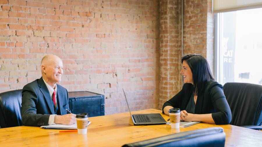 Two people engaged in a discussion at a table with a laptop and coffee cups, perhaps strategizing on how to hire staff, all set against the backdrop of an exposed brick wall.