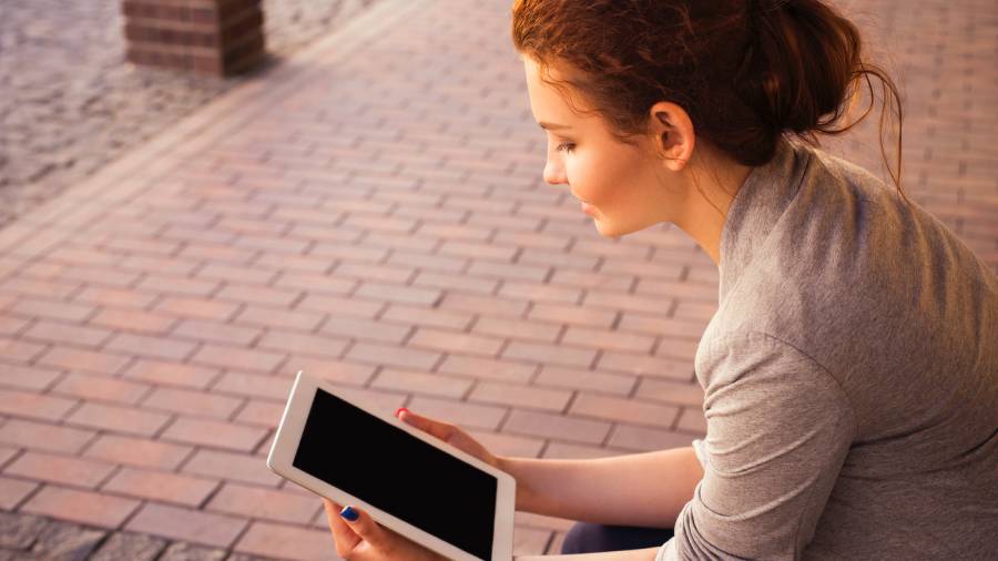 A person with reddish-brown hair, wearing a grey top, is sitting on a brick surface and looking at a tablet displaying Human Resources information on its black screen.