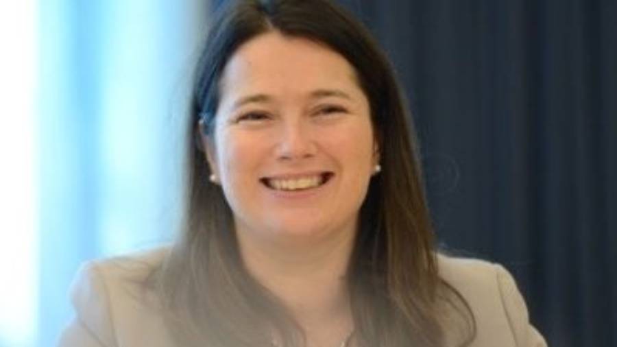 A woman with long dark hair, who works in Human Resources, smiles while wearing a light-colored blazer. She is sitting indoors in front of a dark background.