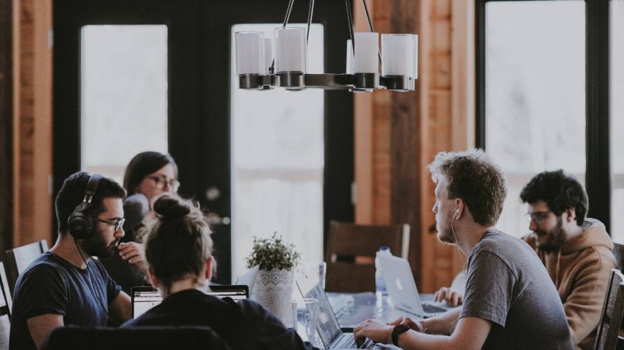 Six people sit around a table in a well-lit room with laptops and notepads, appearing to be in an HR meeting or discussion. A chandelier hangs above the table.