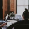 A group of people working on laptops sit around a table in a well-lit room, promoting a healthy work environment. One person wears headphones, likely to focus. A small potted plant adds a touch of nature to the scene, possibly helping alleviate health issues related to poor air quality.