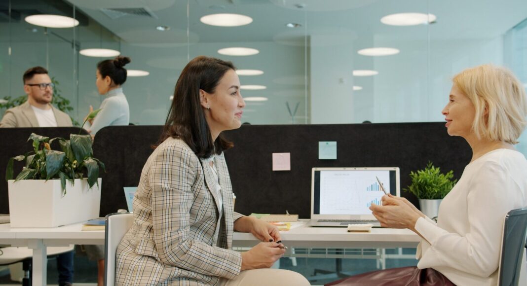 Two women talking at a desk in an office.