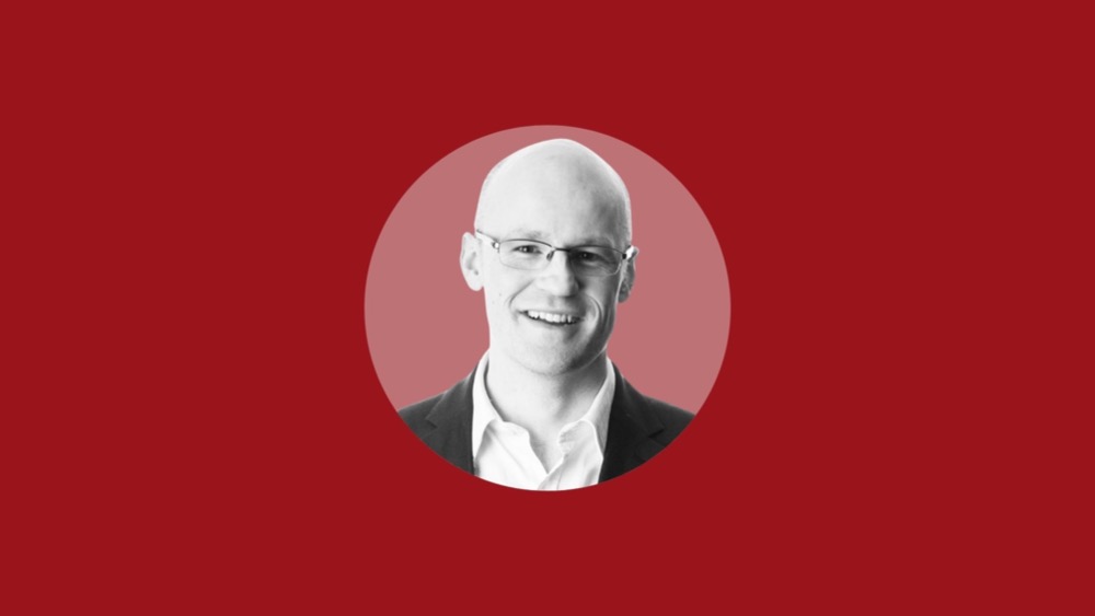 A headshot of a bald man wearing glasses and a suit jacket, smiling confidently against a red background, subtly advocating for addressing the gender pay gap.