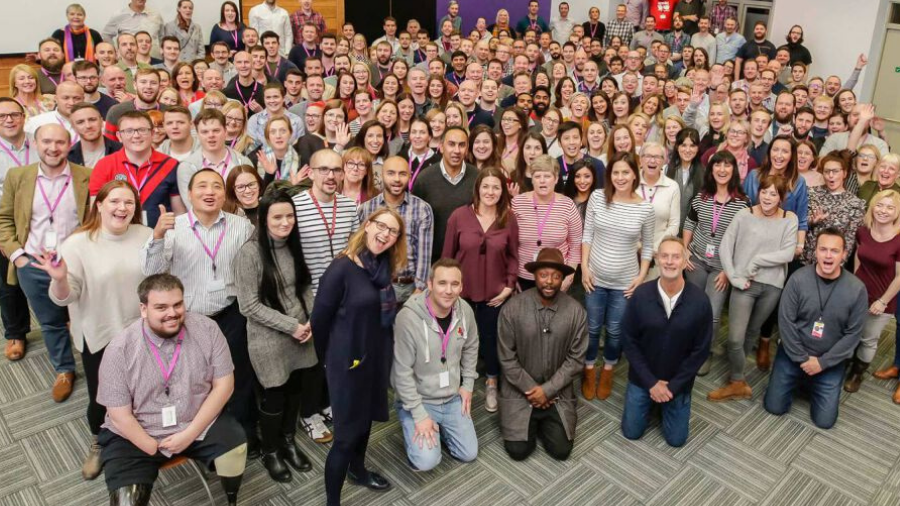 A large group of people, both men and women, pose for a group photo in an indoor setting. Most individuals are smiling, some wearing name badges and lanyards. The mood is especially cheerful as they celebrate the success of their new four day week initiative.