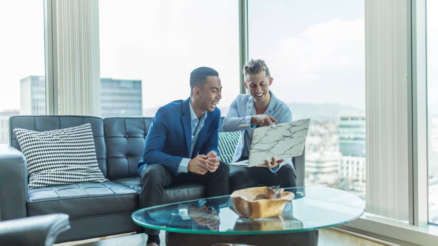 Two men in business attire sit on a black leather couch in a modern office, smiling and looking at a laptop together on a glass coffee table, perhaps discussing pay awards.