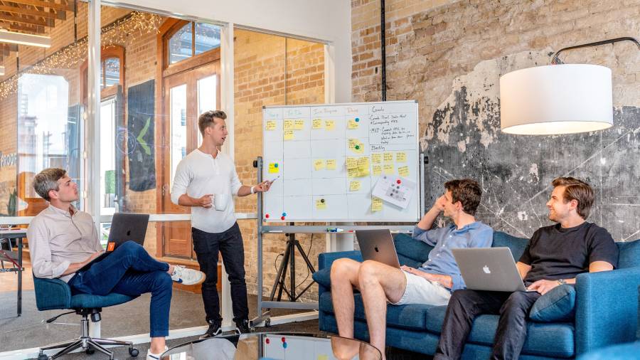 Four people having a casual business meeting in a modern office, one standing and presenting with a whiteboard about how to fill vacancies, while three others sit on chairs and a sofa with laptops.