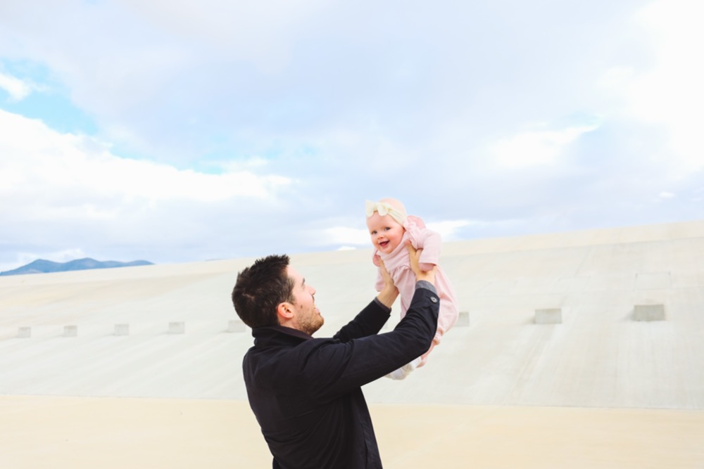 An adult wearing a dark jacket holds a smiling baby dressed in pink with a bow on her head, lifting her up against a bright, cloudy sky backdrop—a heartwarming moment for new fathers.