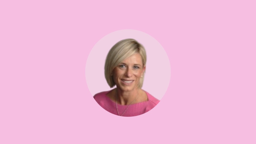 A woman with short blonde hair smiles while wearing a pink sweater, set against a solid pink background, radiating positivity and mental health.